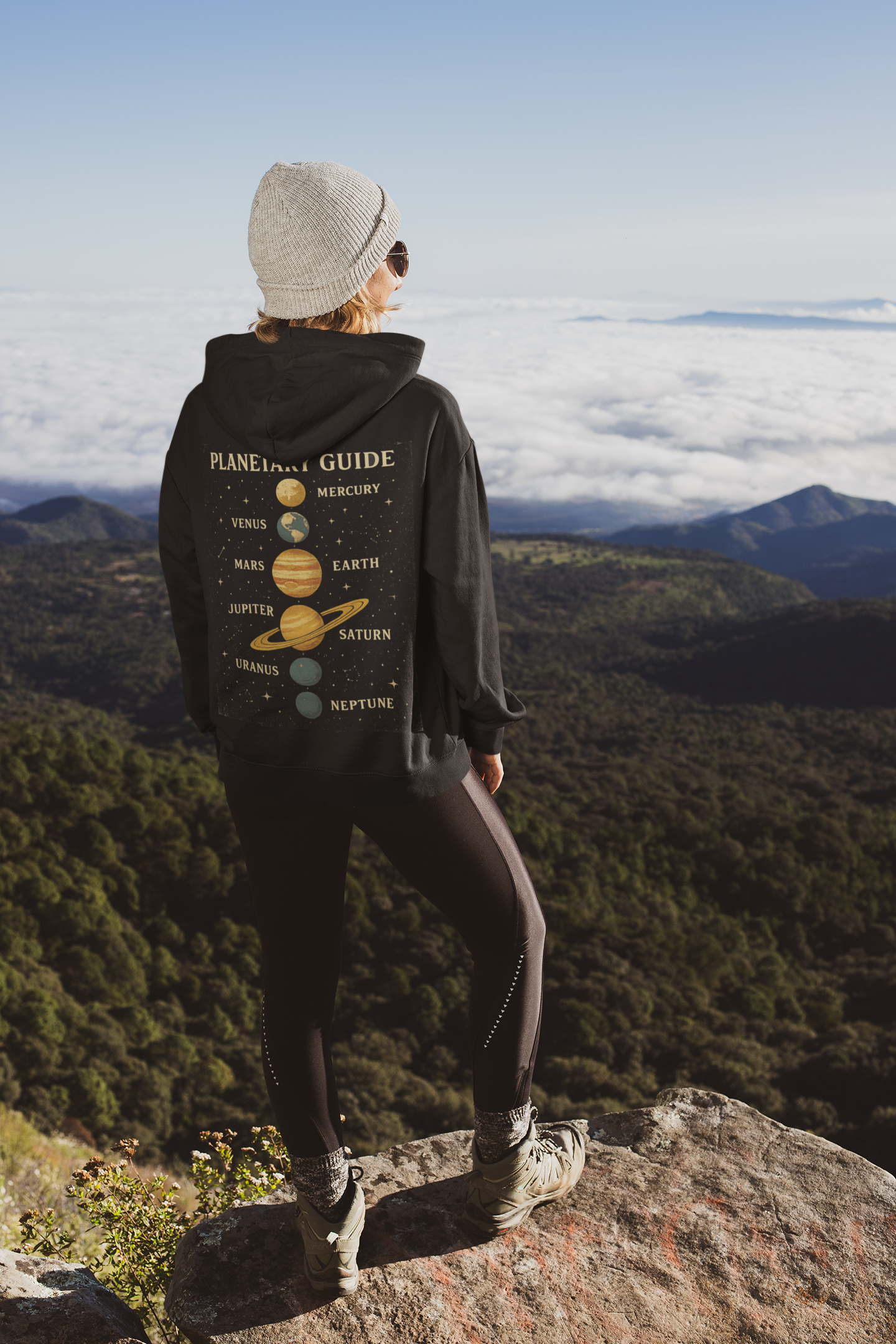 Woman standing on mountain rock wearing black hoodie with solar system planets print on back
